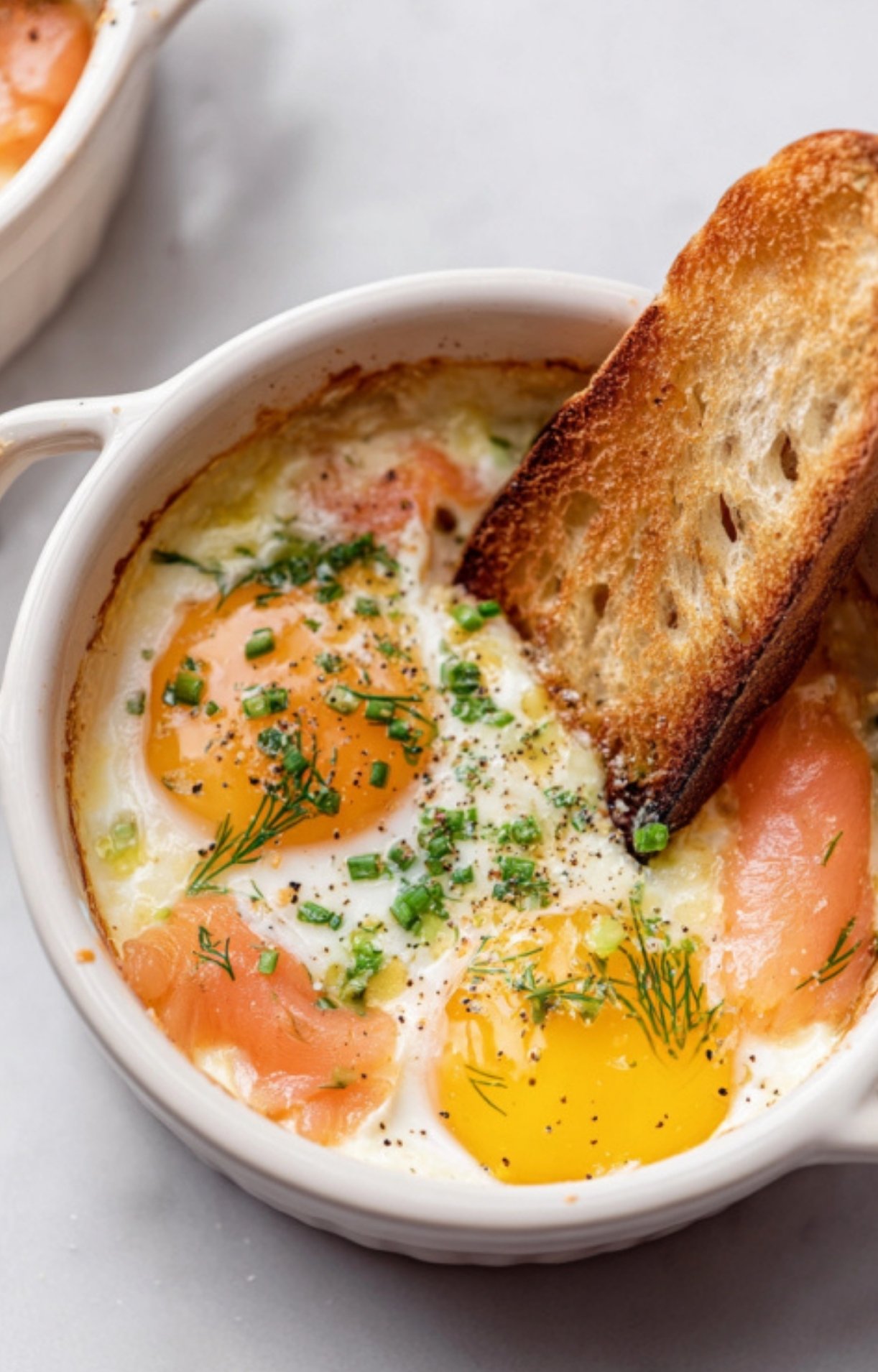 Baked eggs with smoked salmon, fresh dill, and chives in a white ramekin, served with toasted bread for dipping.