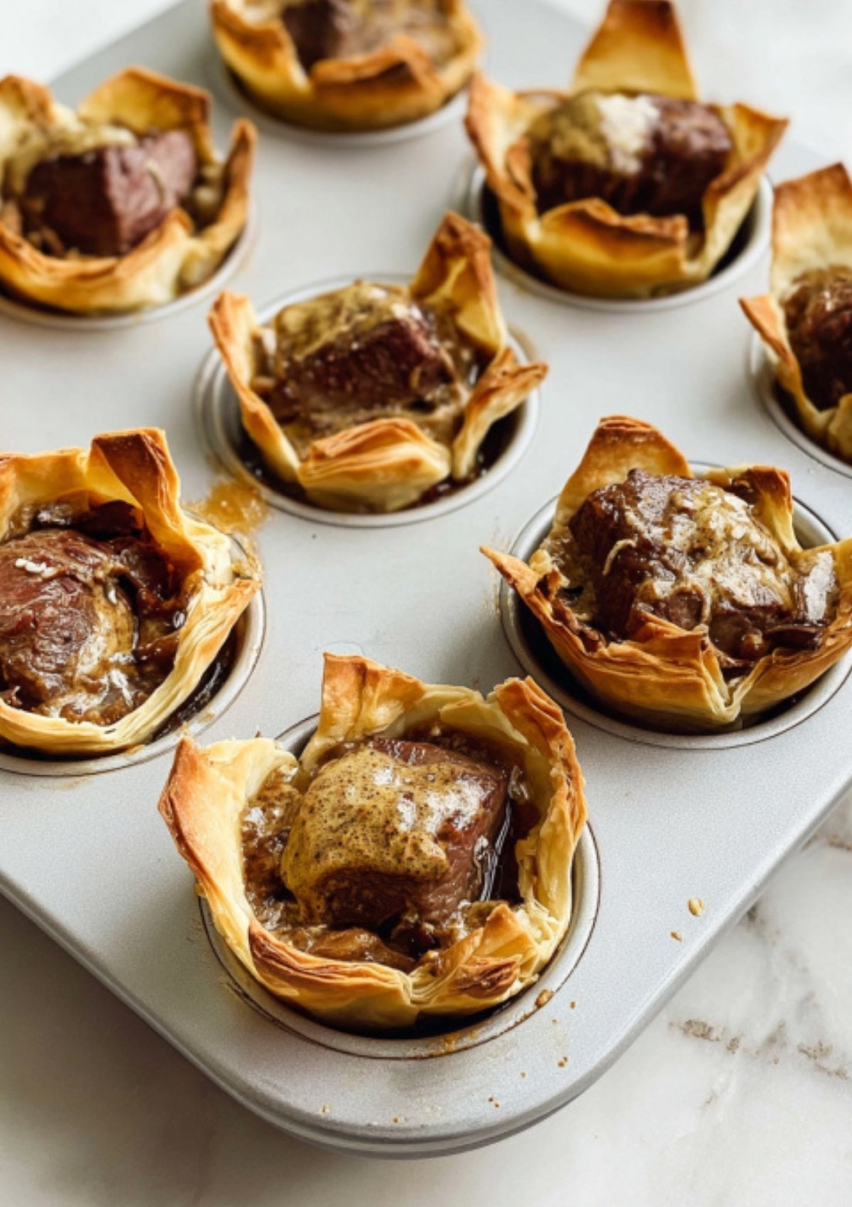 Golden mini beef wellington cups baked in a muffin tin with puff pastry, beef, and mushroom filling, perfect for holiday appetizers or party snacks.