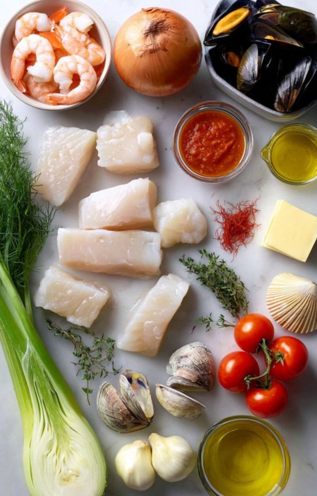 Flat lay of fresh ingredients for bouillabaisse: white fish, shrimp, mussels, clams, fennel, leeks, onion, tomatoes, herbs, olive oil, butter, garlic, saffron. Perfect for easy, impressive Mediterranean meals.