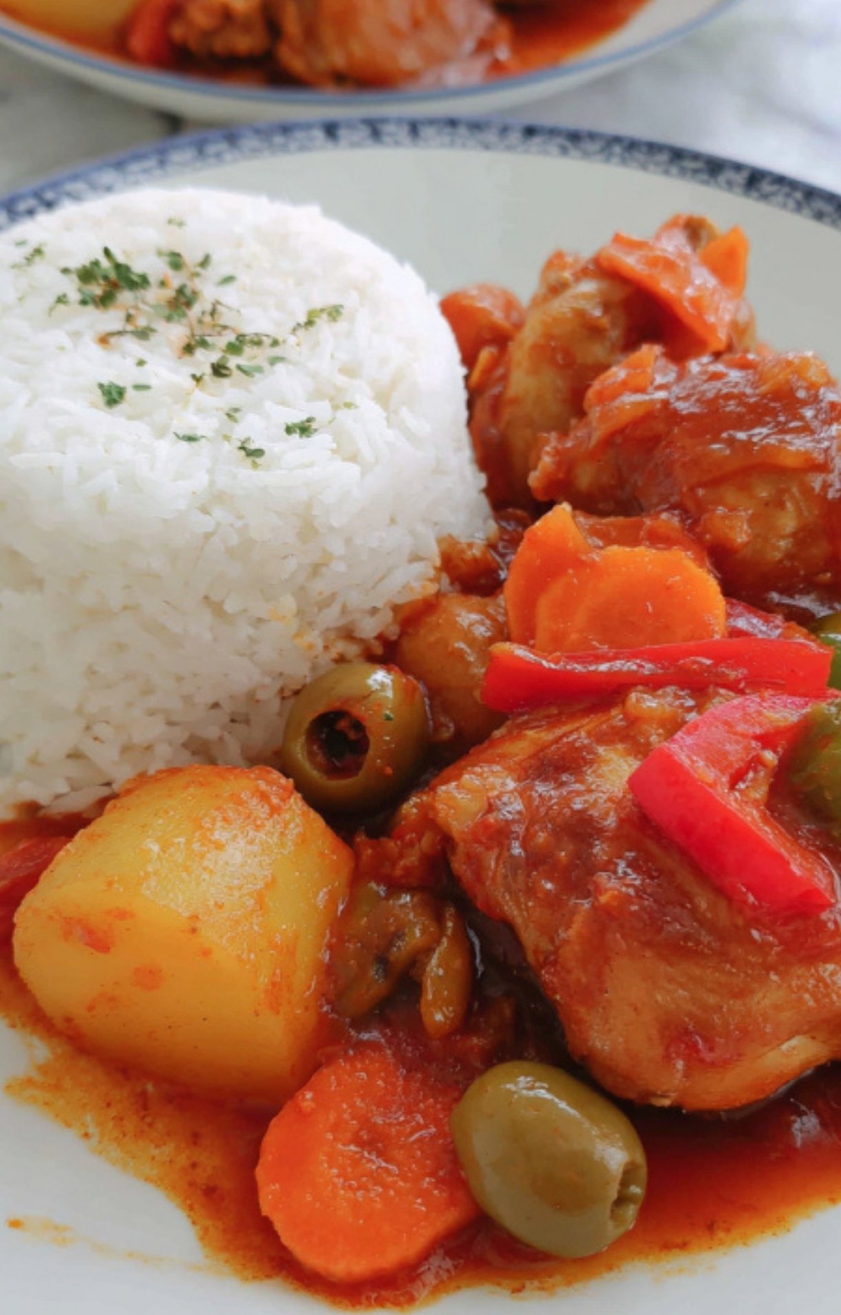 A plate with fluffy white rice topped with herbs and a generous serving of chicken caldereta with carrots, potatoes, bell peppers, and green olives in tomato sauce. Classic Filipino stew for family meals.
