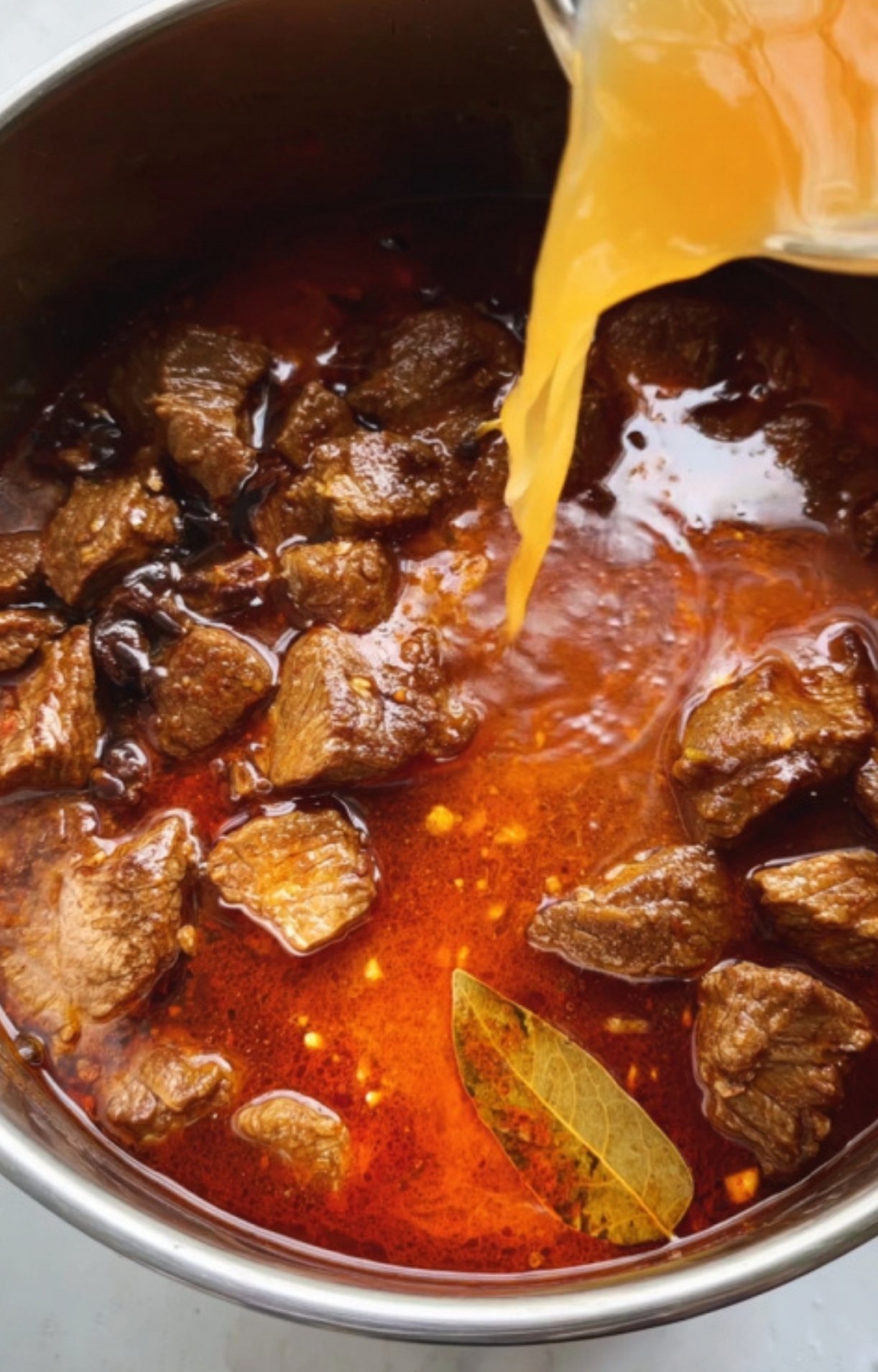 Pouring golden broth into a pot of browned beef cubes with bay leaf, a key step for juicy and flavorful German goulash. Great for family comfort food nights.