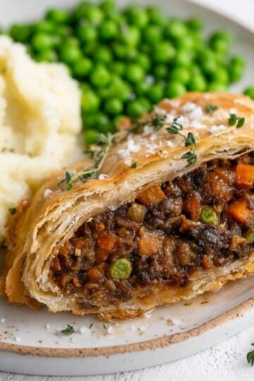 A hearty vegan beef Wellington sliced open, showing a savory mushroom and veggie filling inside golden puff pastry, served with mashed potatoes and peas. Ideal for vegan Christmas dinner or Thanksgiving.