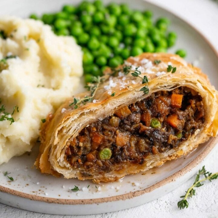 A hearty vegan beef Wellington sliced open, showing a savory mushroom and veggie filling inside golden puff pastry, served with mashed potatoes and peas. Ideal for vegan Christmas dinner or Thanksgiving.