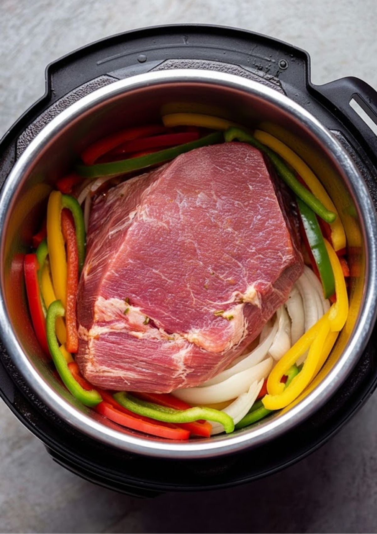 Uncooked corned beef brisket with sliced bell peppers and onions in a pressure cooker, ready for seasoning and broth. Easy start to homemade corned beef.