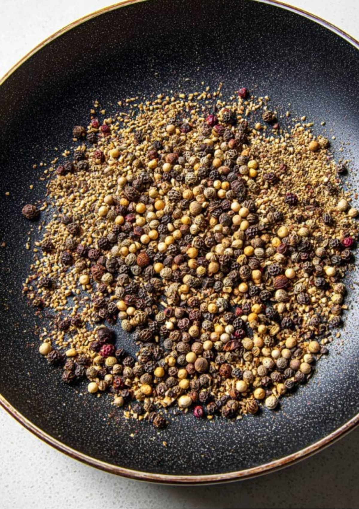 Whole pickling spices including mustard seeds, peppercorns, and coriander seeds toasting in a black skillet.