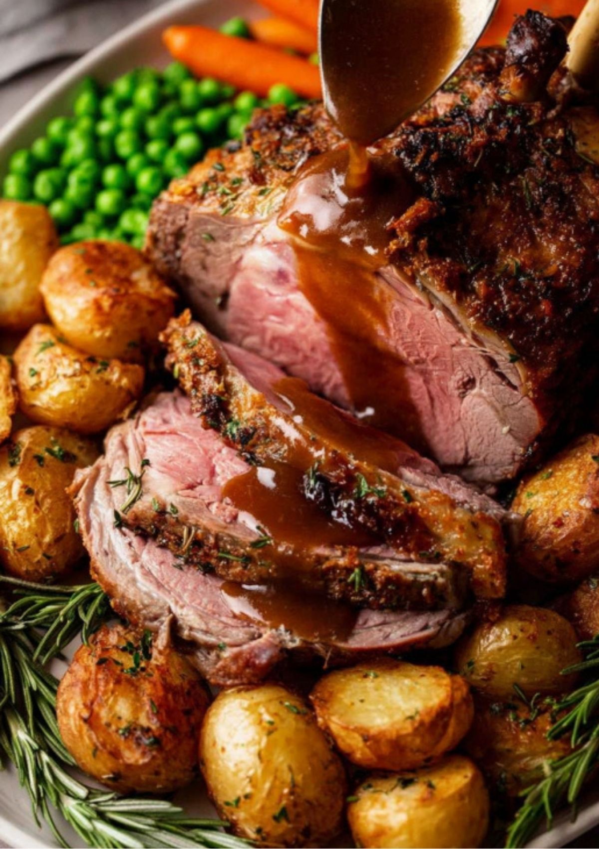Sliced roast leg of lamb with garlic rosemary crust, topped with rich gravy and served with roast potatoes, peas, and carrots