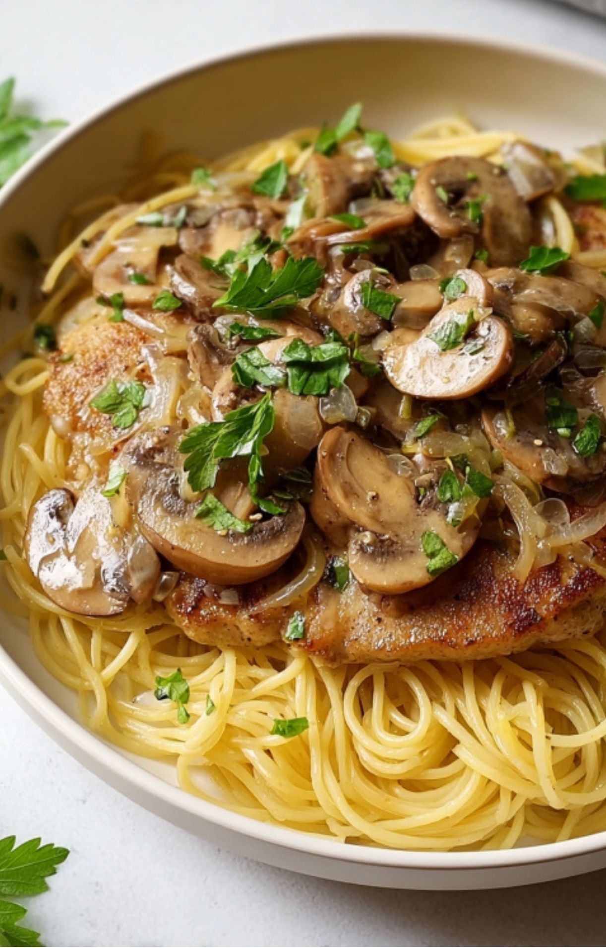 Bowl of veal scallopini with mushrooms and parsley served on top of a bed of cooked spaghetti.