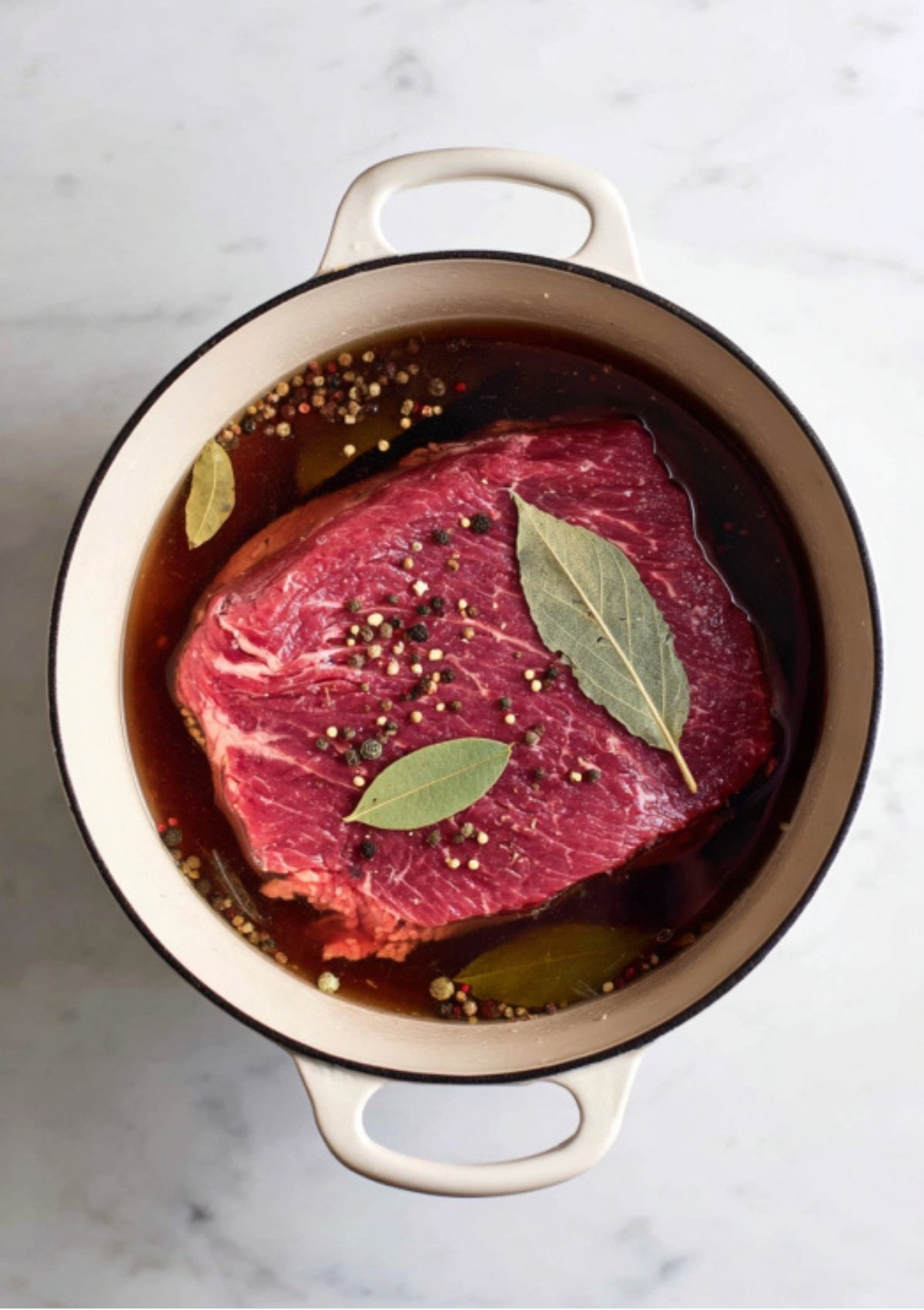 Corned beef brisket submerged in broth with bay leaves and peppercorns in a Dutch oven, starting the classic Irish recipe.