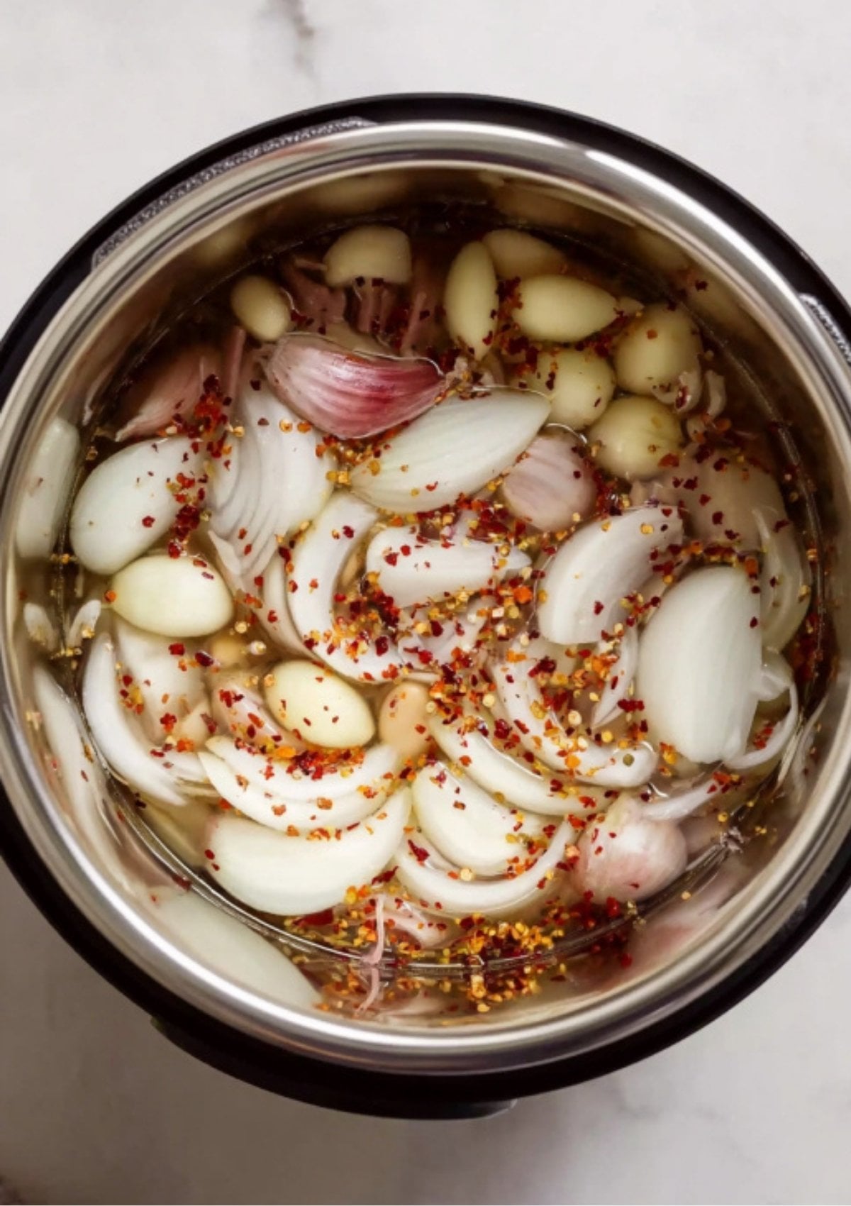 Onions, garlic cloves, and red pepper flakes simmering in liquid inside the cooker to build a flavorful base for Instant Pot Corned Beef.