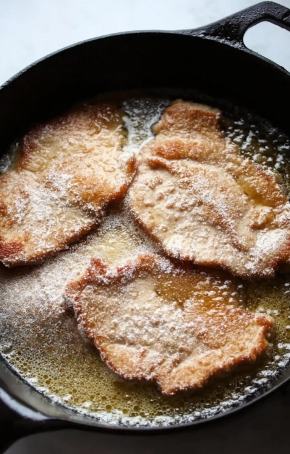 Three breaded veal cutlets sizzling in a cast-iron skillet with hot oil.