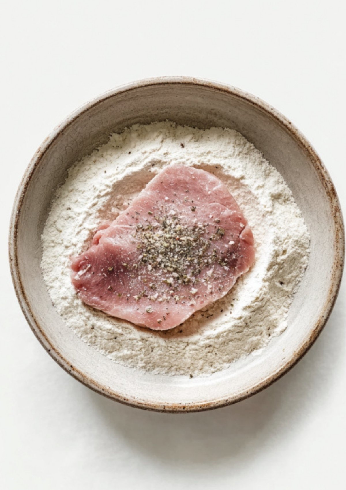 Raw veal scallopini cutlet in a bowl of seasoned flour, ready for pan-searing.