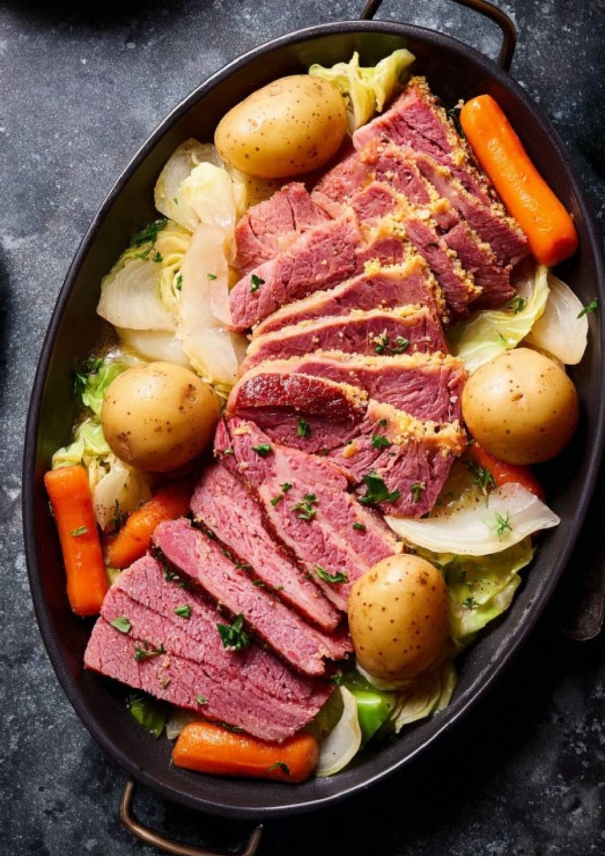 Oval serving dish with sliced corned beef, boiled potatoes, carrots, cabbage, and onions, garnished with fresh herbs for a traditional meal.