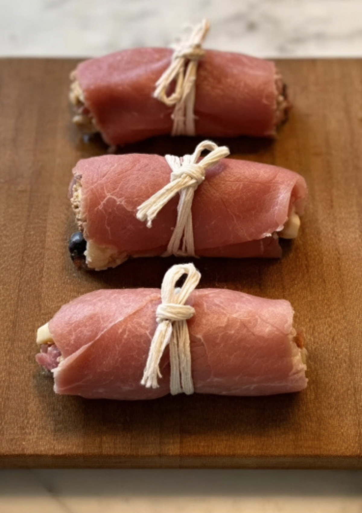 Three braciole rolls tied with kitchen twine, resting on a wooden board before browning.