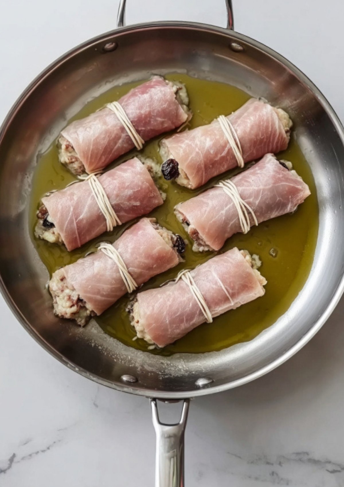 Uncooked braciole rolls wrapped in prosciutto and tied with string, sitting in olive oil in a skillet before browning.