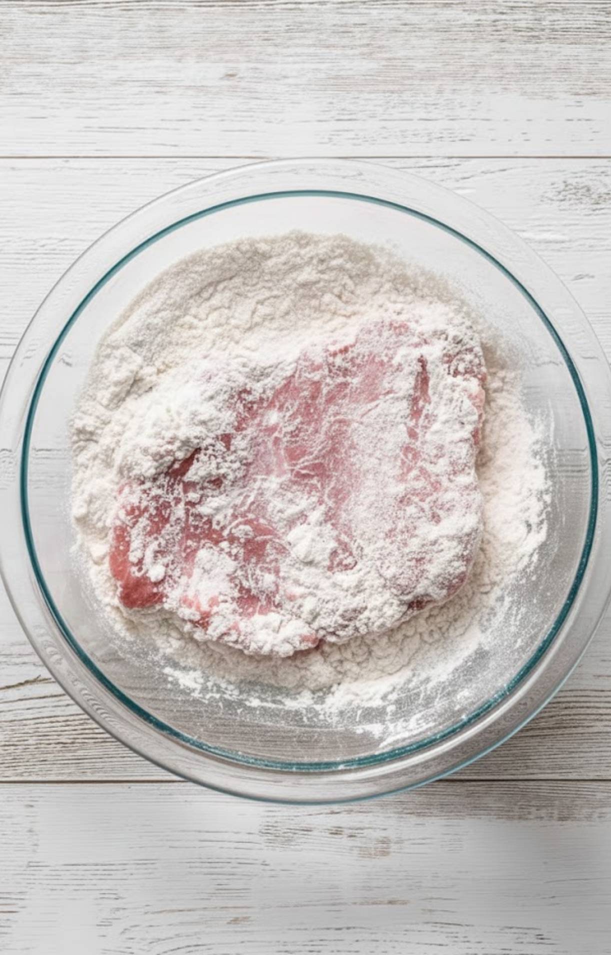 Raw veal cutlet coated in flour in a glass bowl, the first step to making crispy, tender veal scallopini with mushrooms.