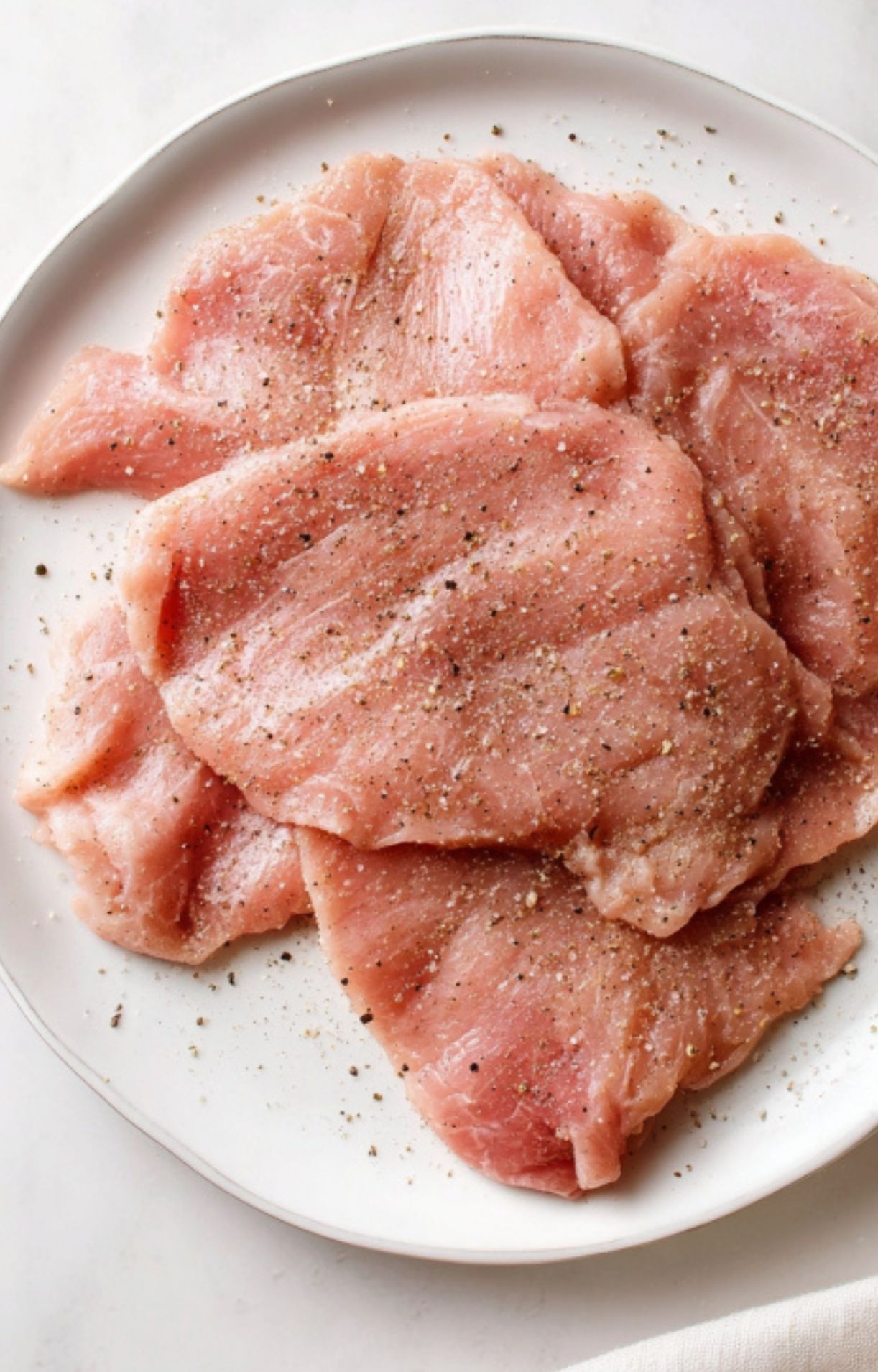 Thin veal cutlets sprinkled with salt and black pepper on a white plate, first step for making veal Parmesan