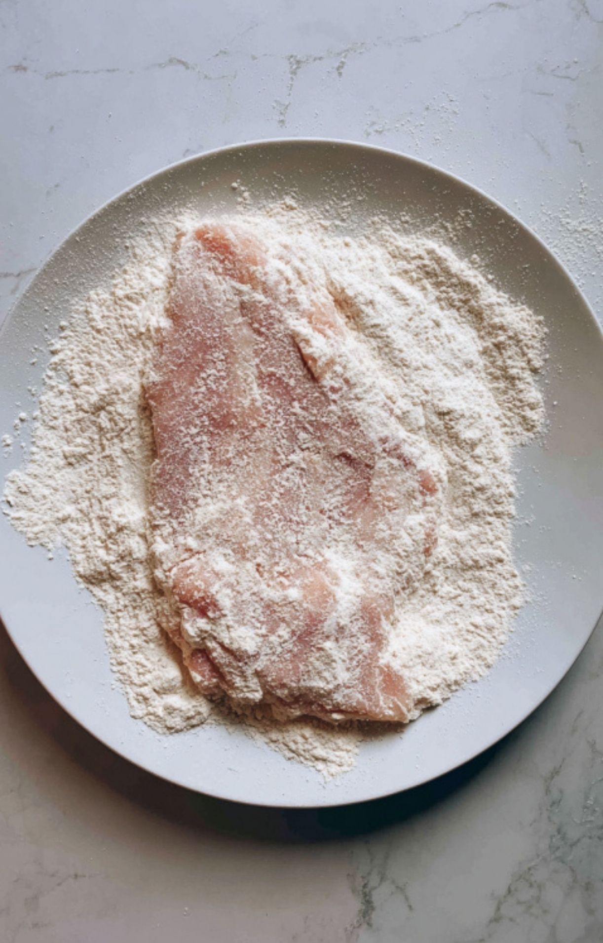 Thin veal cutlet coated in flour on a white plate, ready for pan-searing. Essential first step for making veal scallopini with mushroom sauce.