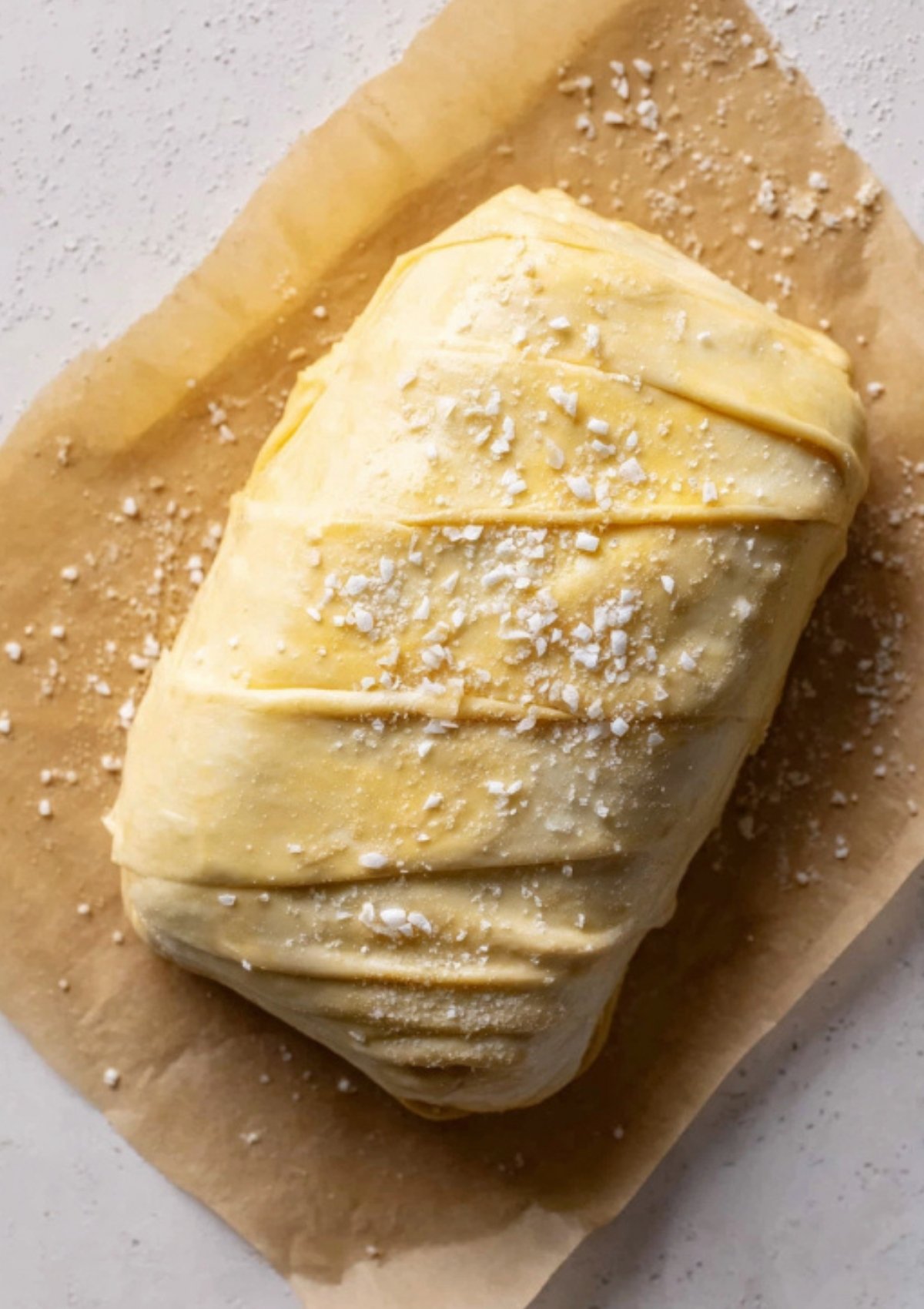 Unbaked Beef Wellington wrapped in pastry, sprinkled with sea salt and brushed with egg wash, sitting on parchment paper. Perfect step for preparing this elegant holiday dish.