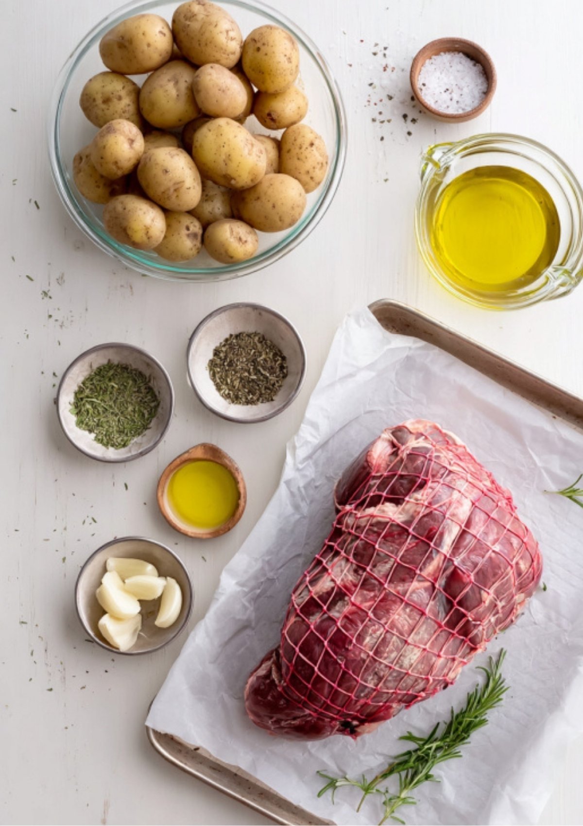 Overhead shot of raw lamb in netting, baby potatoes, olive oil, garlic, and herbs arranged on a white surface