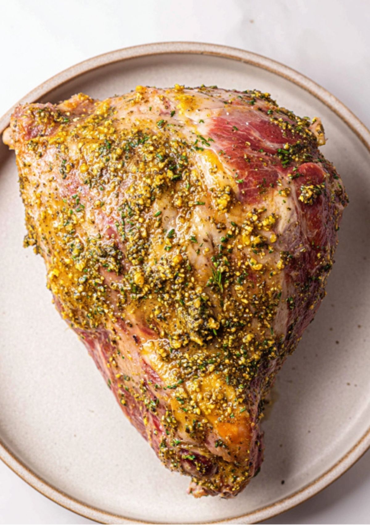 Uncooked leg of lamb generously coated with mustard, garlic, and herb marinade, displayed on a plate.