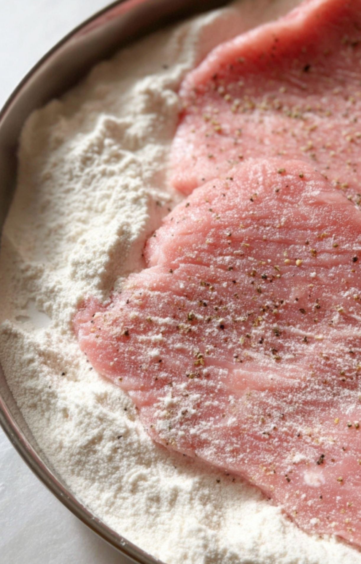 Raw veal cutlets being coated in seasoned flour in a shallow pan, ready for scallopini.
