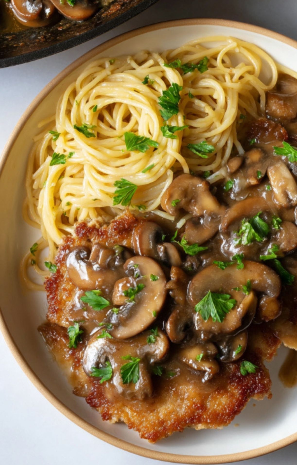 Plate with spaghetti and a veal cutlet topped with mushroom sauce and sprinkled with fresh parsley.