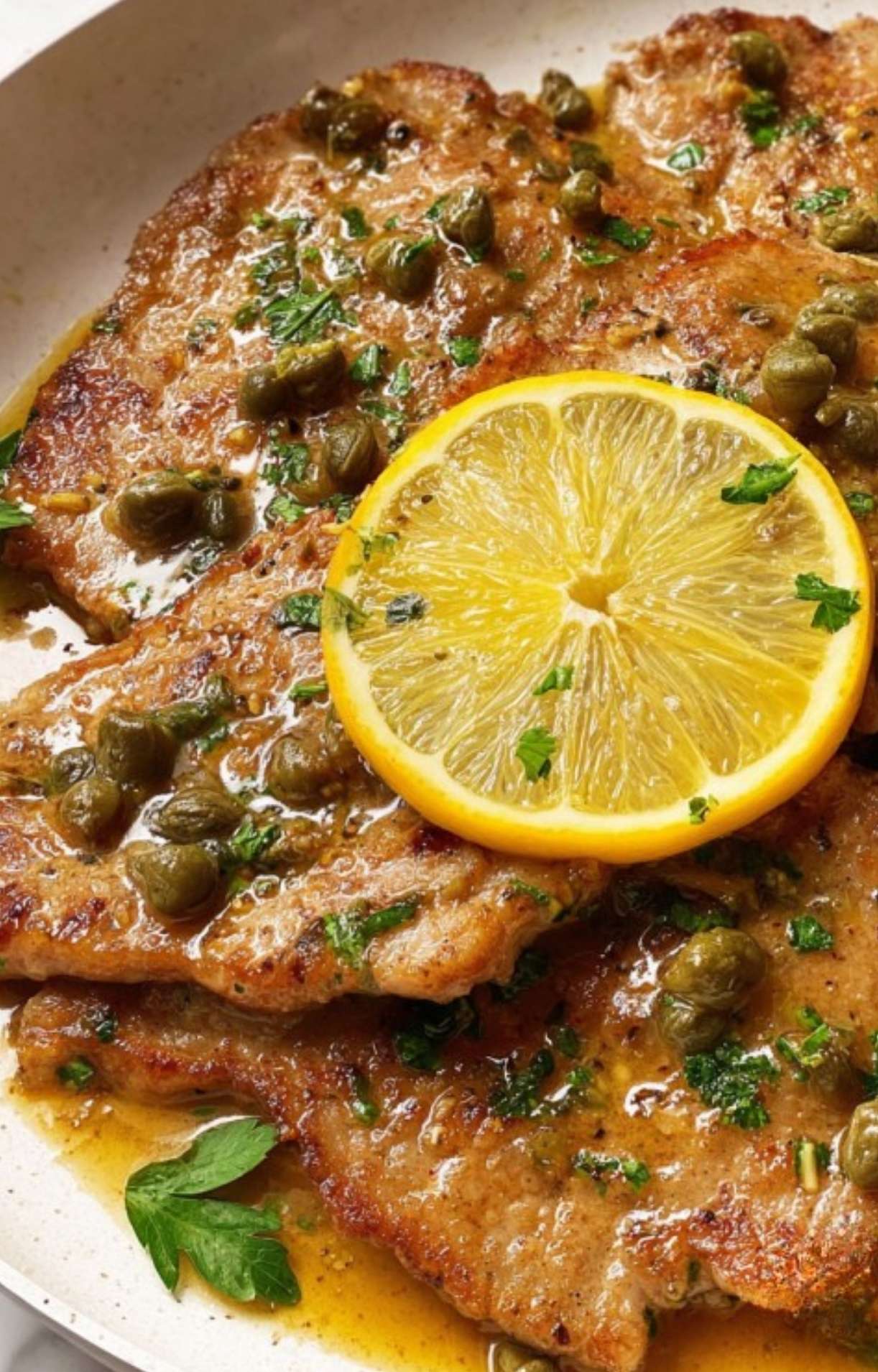 Veal scallopini in a pan with golden lemon caper sauce and fresh lemon slices.