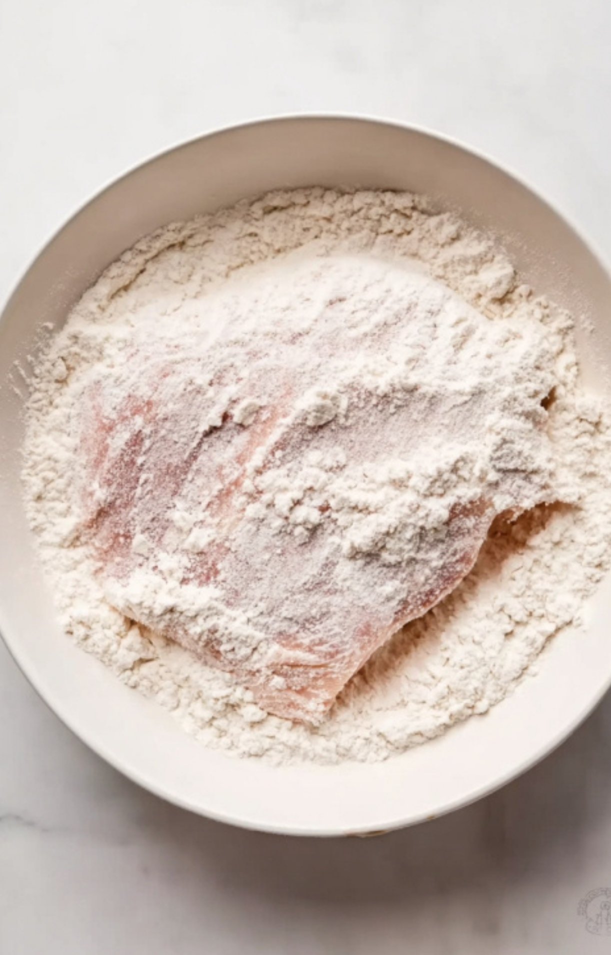 Raw veal cutlet being coated in a bowl of flour, prepping for crispy breaded veal Parmesan