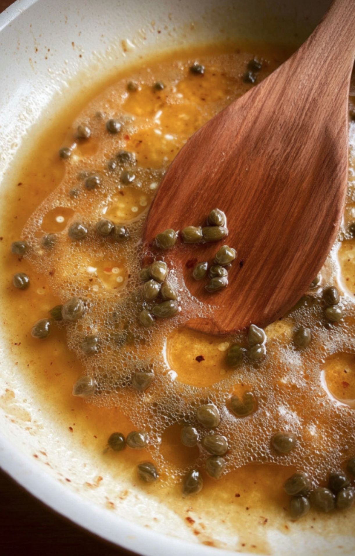 A wooden spatula stirring capers and a golden buttery liquid in a pan to create the base for Veal Scallopini in Lemon Sauce.