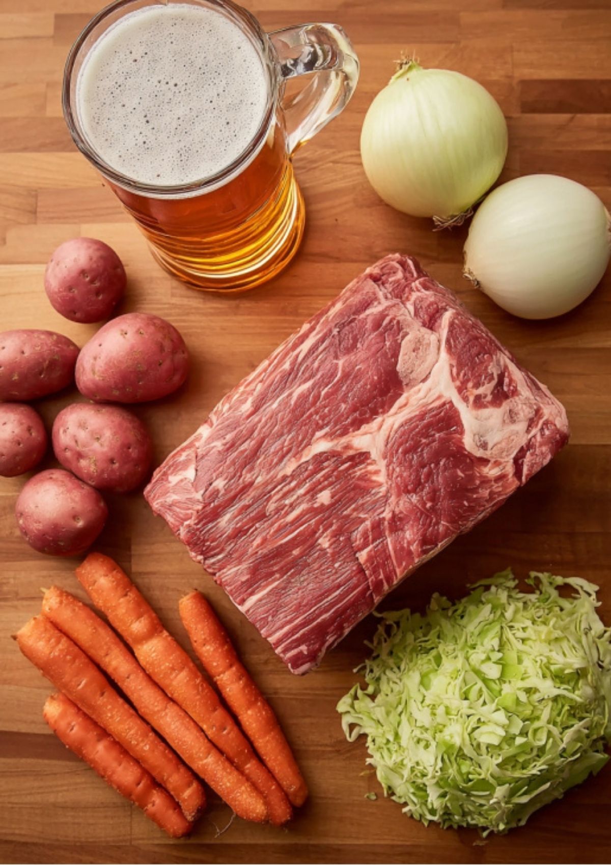 Raw corned beef brisket, beer, onions, red potatoes, carrots, and chopped cabbage on a wooden surface. All the essentials for an easy Instant Pot dinner.
