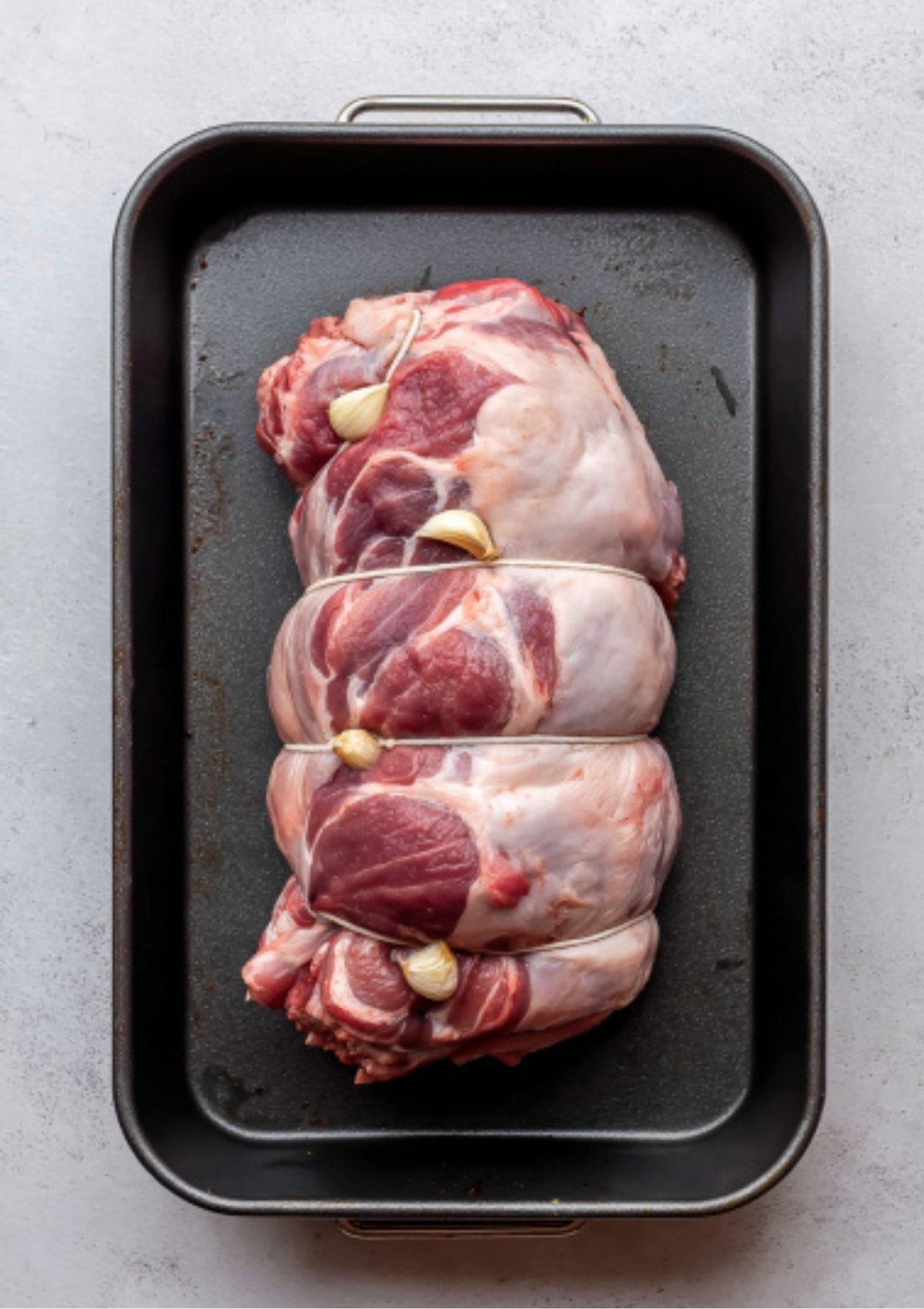 Raw boneless leg of lamb tied with garlic cloves tucked inside, in a black roasting pan before seasoning