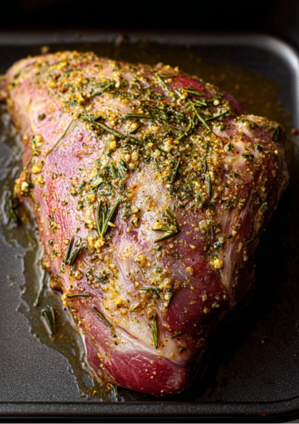 Raw leg of lamb coated with a garlic, rosemary, and mustard marinade in a roasting pan before cooking.