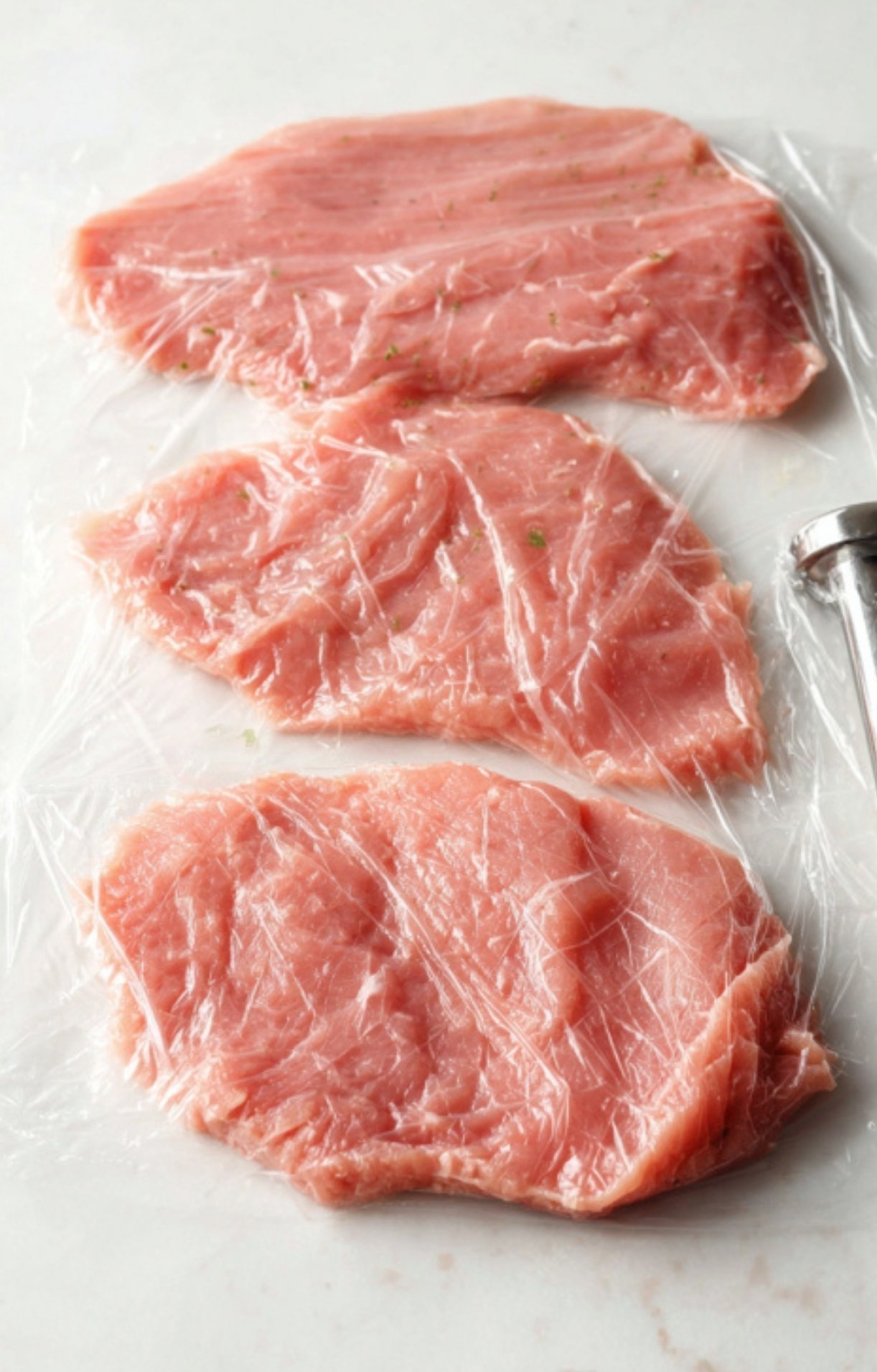 Three raw veal cutlets covered in plastic wrap, pounded thin and ready for cooking.