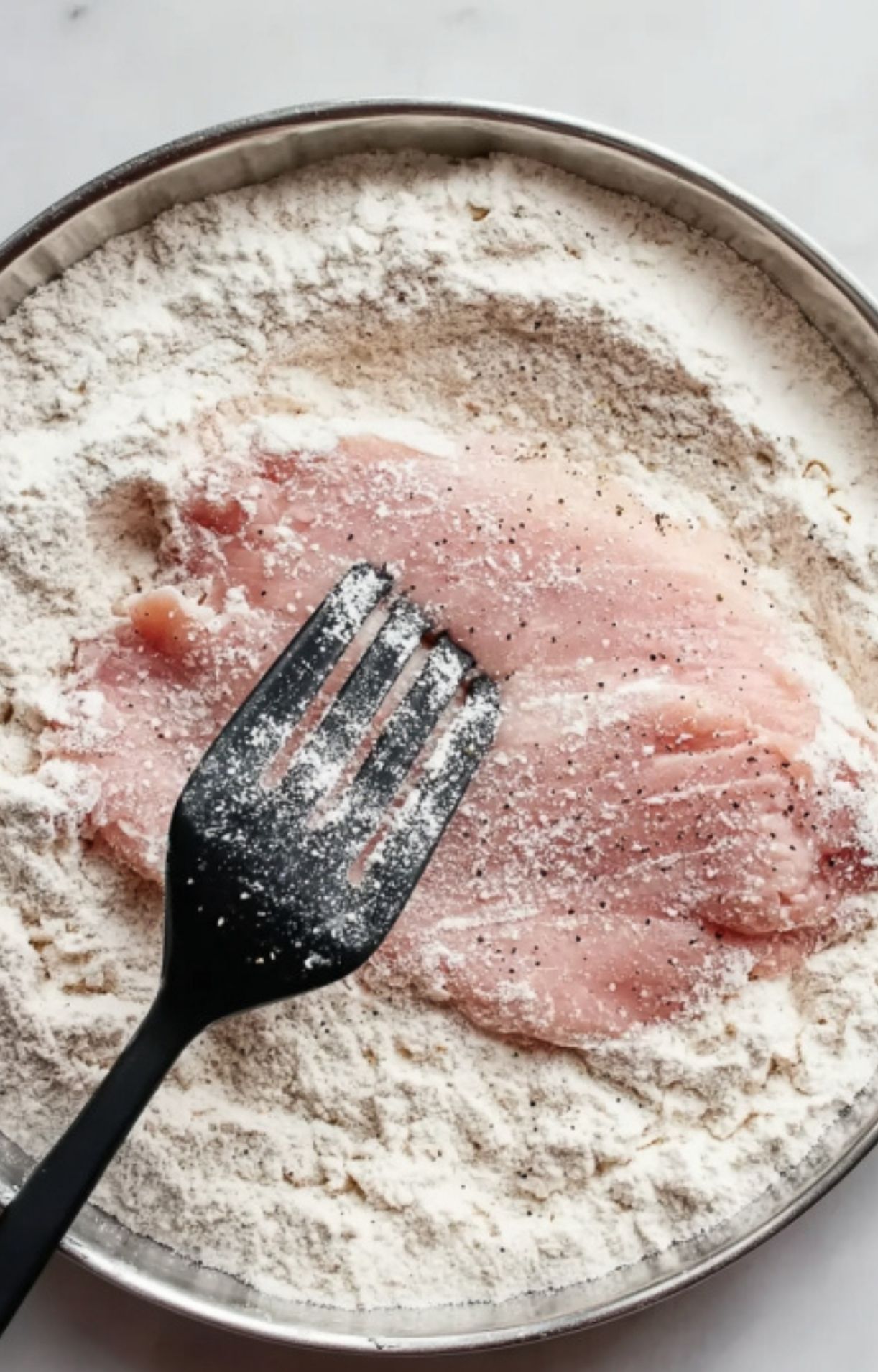 A hand using a fork to coat a piece of raw veal in a shallow bowl of white flour.