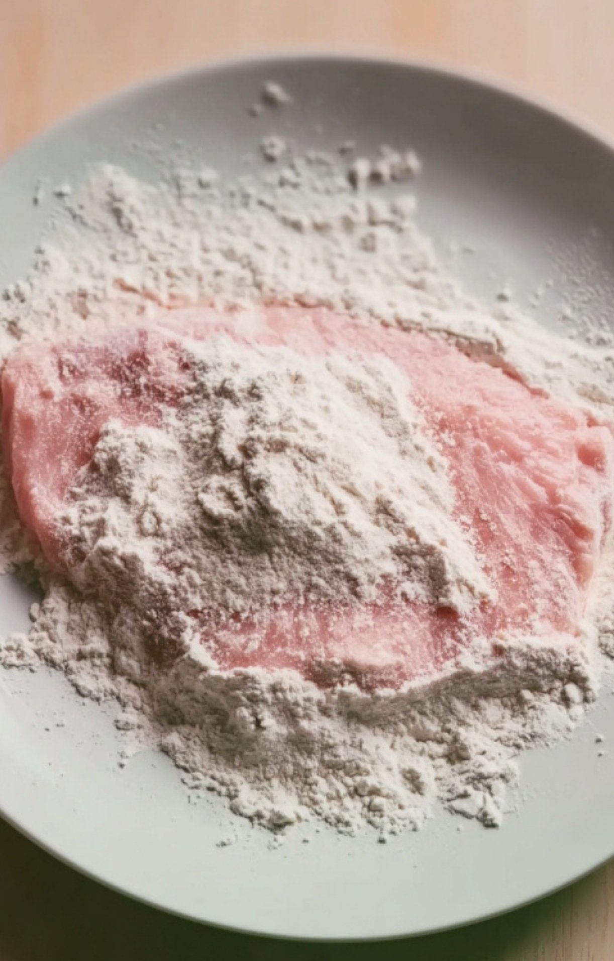 A raw veal cutlet being coated in white flour on a plate as the first step for Veal Scallopini in Lemon Sauce.