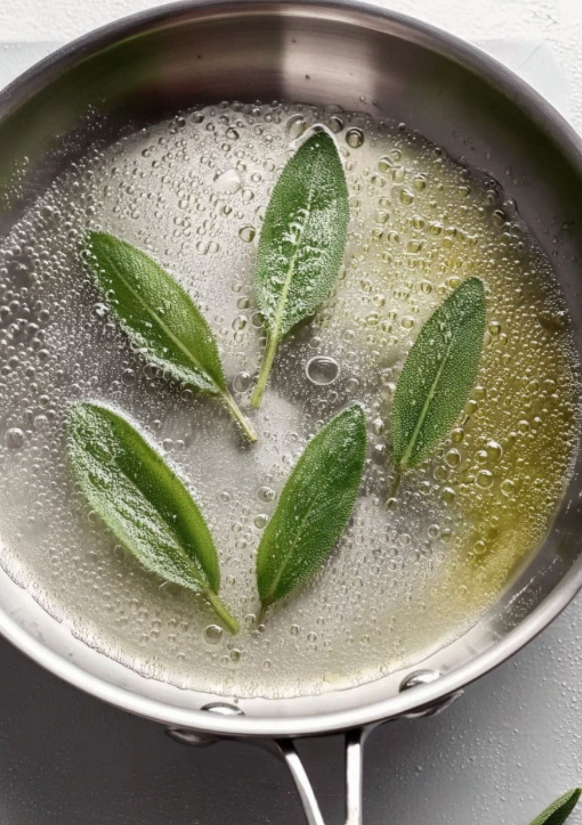 Fresh sage leaves simmering in a bubbling white wine and butter reduction in a silver pan.