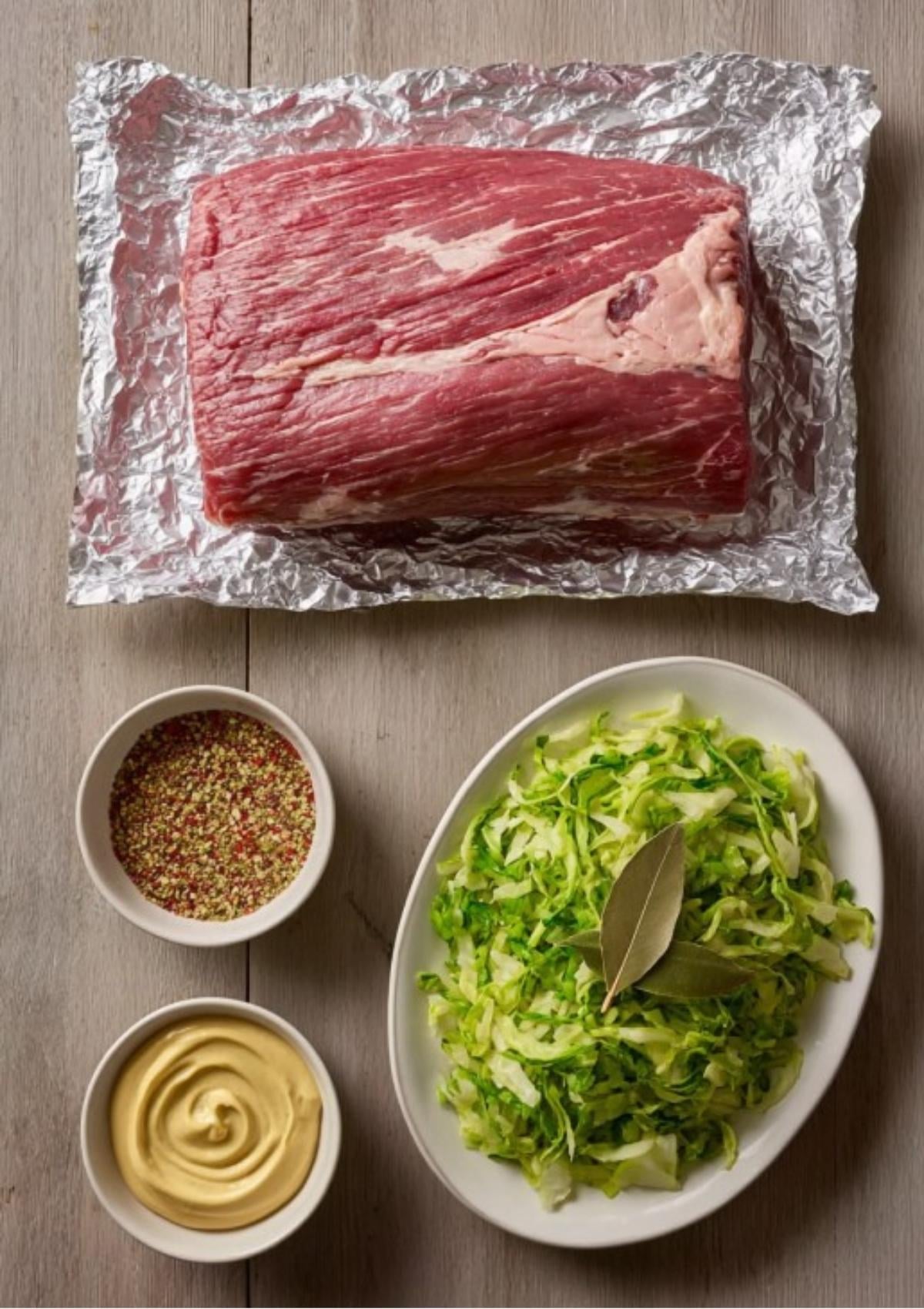 Raw corned beef brisket on foil with bowls of mustard, pickling spice, shredded cabbage, and bay leaves. All the essentials for a perfect baked corned beef dinner.