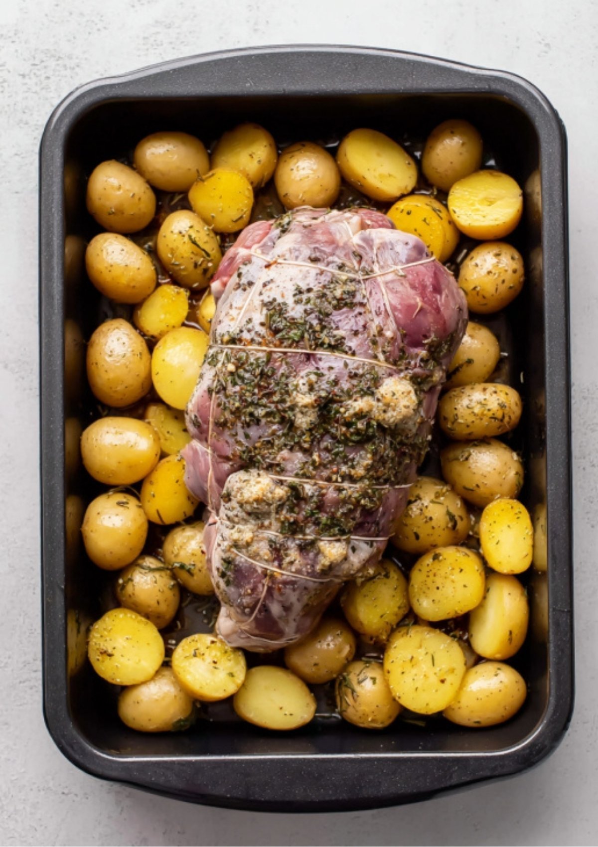 Raw boneless leg of lamb tied with twine and coated with herb and garlic rub, placed on top of small potatoes in a roasting pan