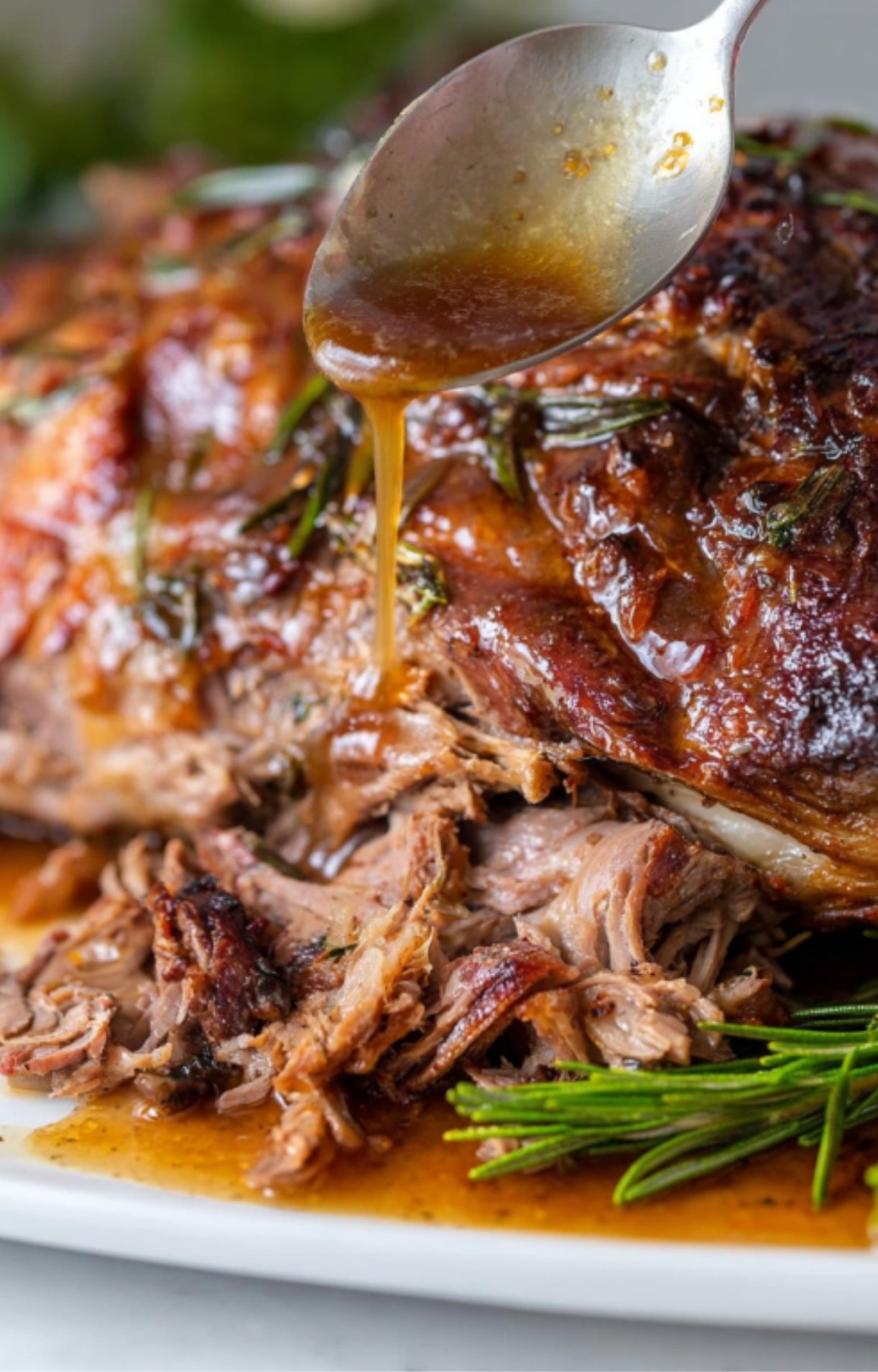 Tender slow roasted leg of lamb on a platter, spoon pouring golden pan juices over shredded meat with fresh rosemary