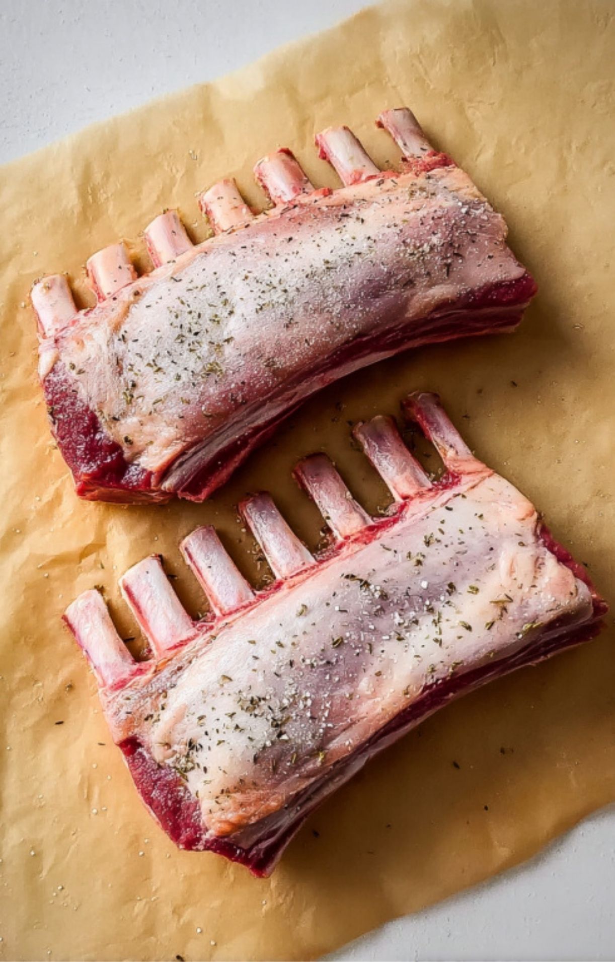Raw racks of lamb seasoned with salt, pepper, and herbs on parchment paper, ready for cooking