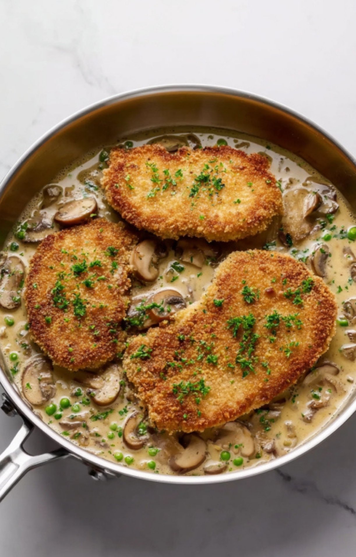 Breaded veal cutlets simmering in a creamy mushroom and pea sauce inside a skillet, garnished with parsley.