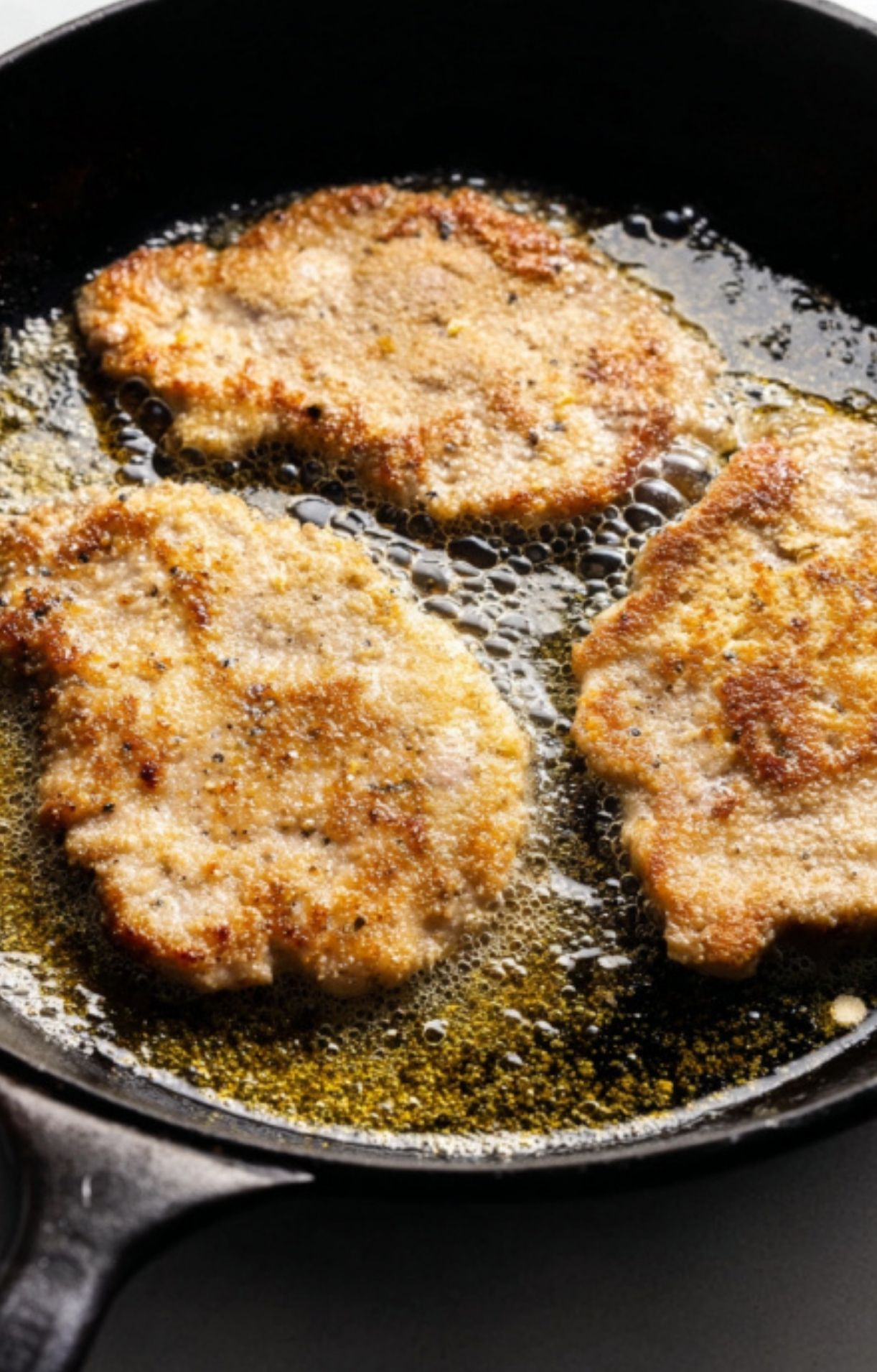 Breaded veal cutlets sizzling in hot oil in a black skillet, turning golden brown.