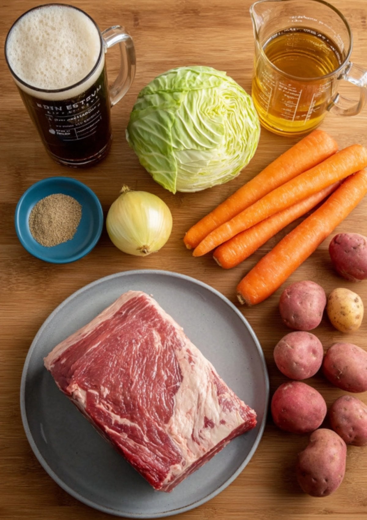 Ingredients for Dutch oven corned beef on a wooden board: brisket, cabbage, carrots, potatoes, onion, mug of beer, broth, and spices. Everything for a classic Irish feast.