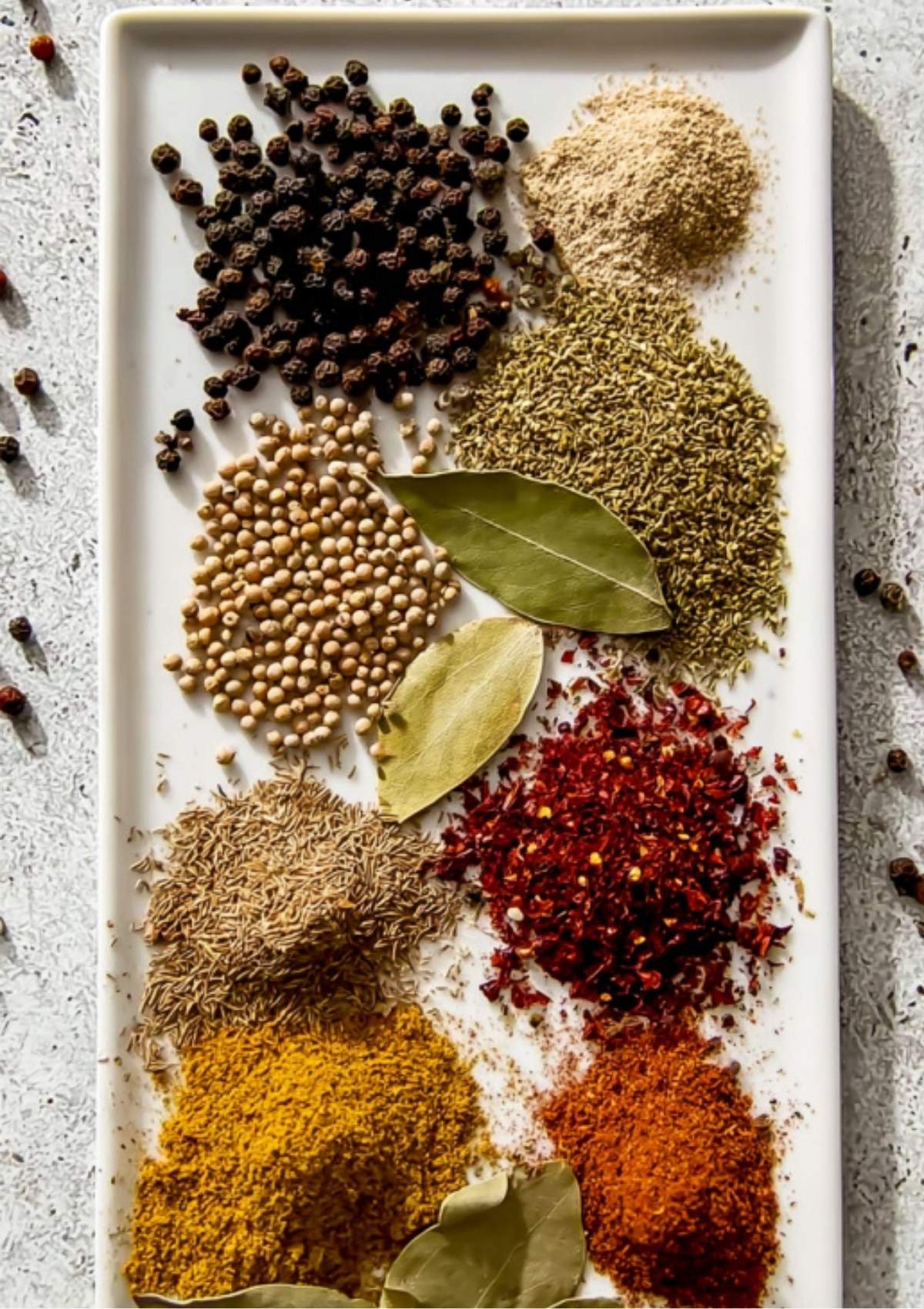 A white tray with small piles of spices: black peppercorns, coriander seeds, bay leaves, crushed red pepper, cumin, and more for homemade corned beef seasoning.