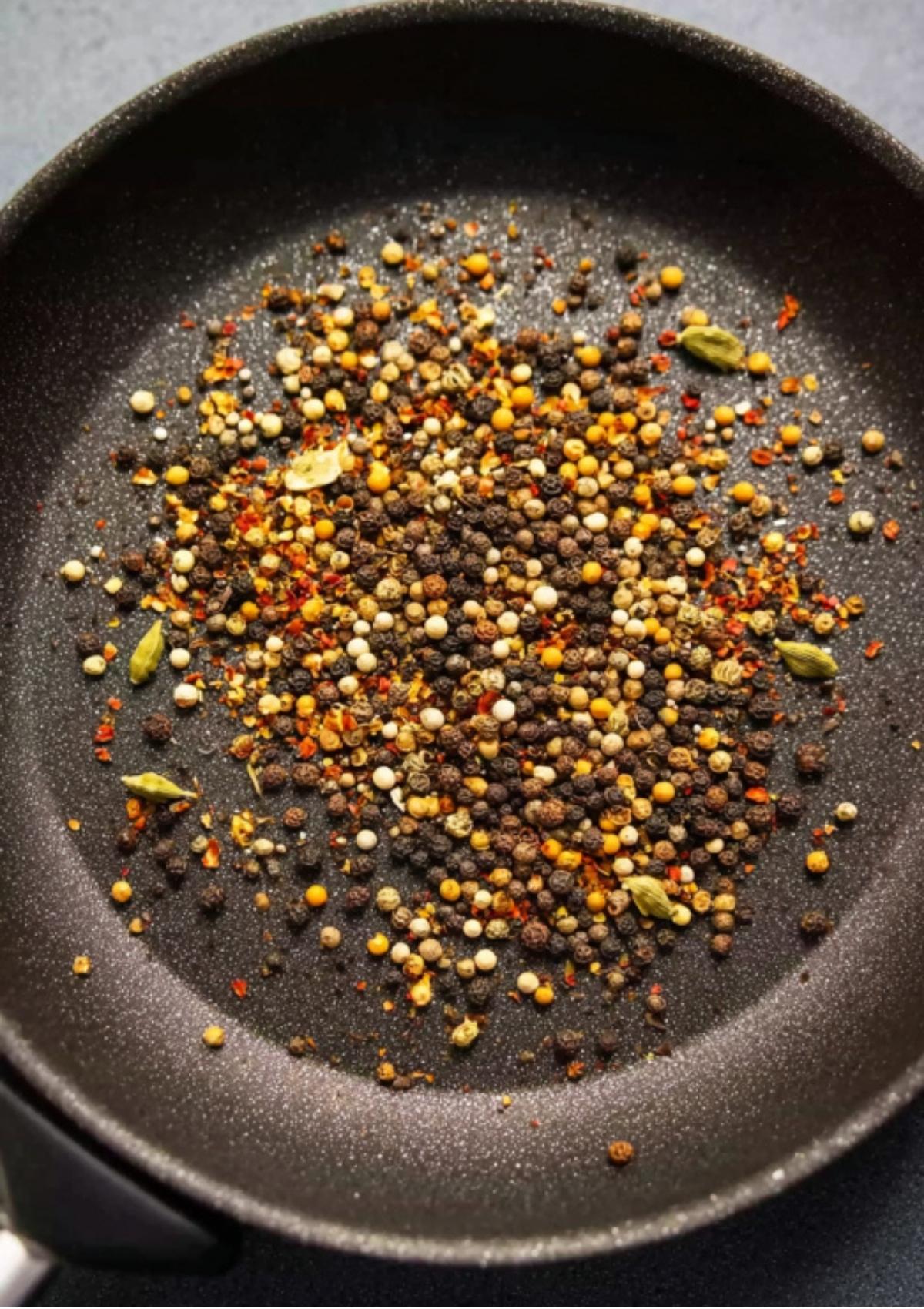 Assorted whole spices like peppercorns, mustard seeds, and cardamom toasting in a black skillet to bring out their flavors for the corned beef brine.