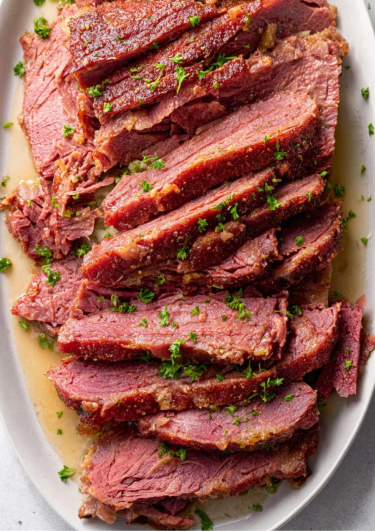 Tender slices of oven baked corned beef with fresh parsley on a serving platter. The ultimate easy recipe for weeknight meals or festive gatherings.