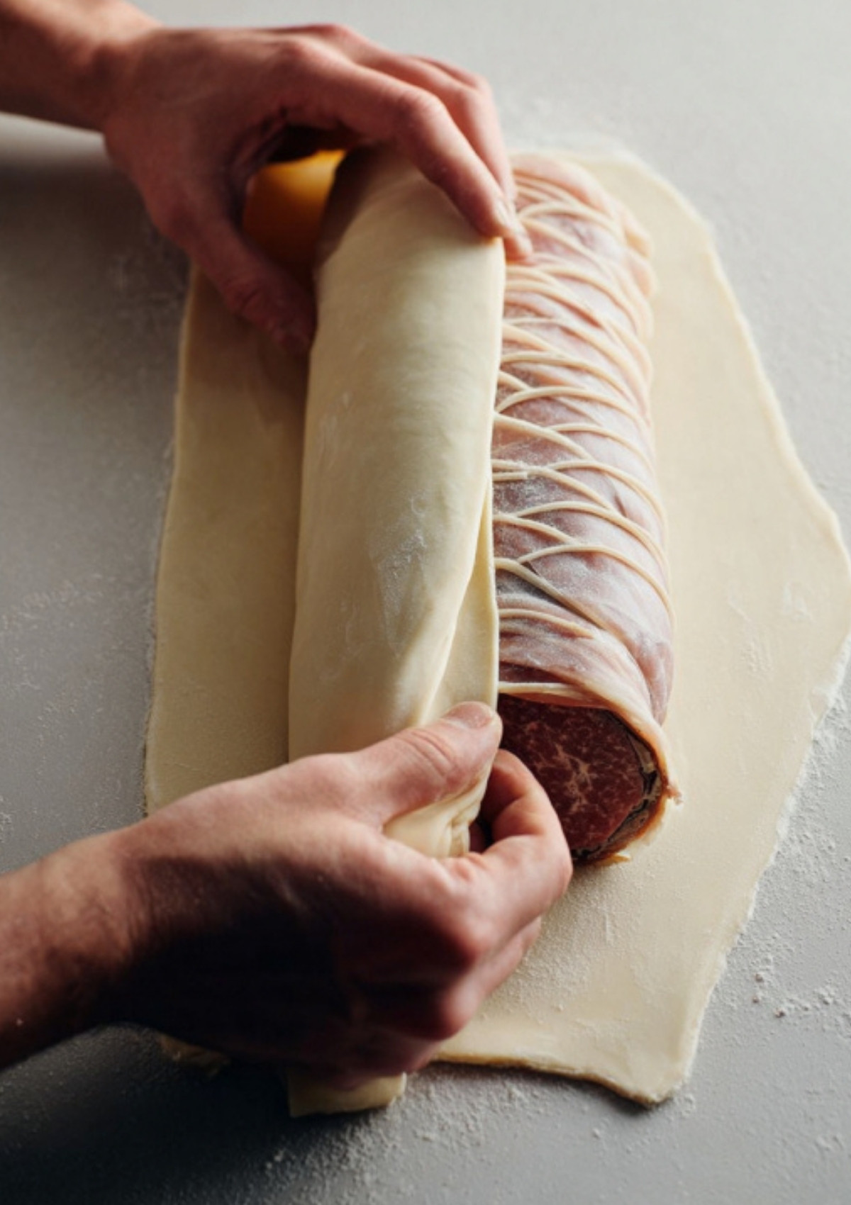 Hands rolling beef Wellington in a sheet of puff pastry, an essential step for this classic holiday dinner recipe.
