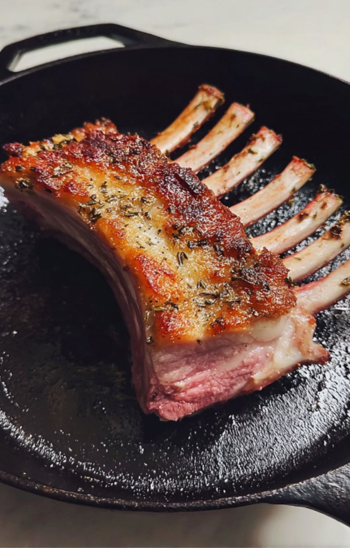 Rack of lamb being seared in a cast iron skillet, developing a golden brown crust before adding herbs