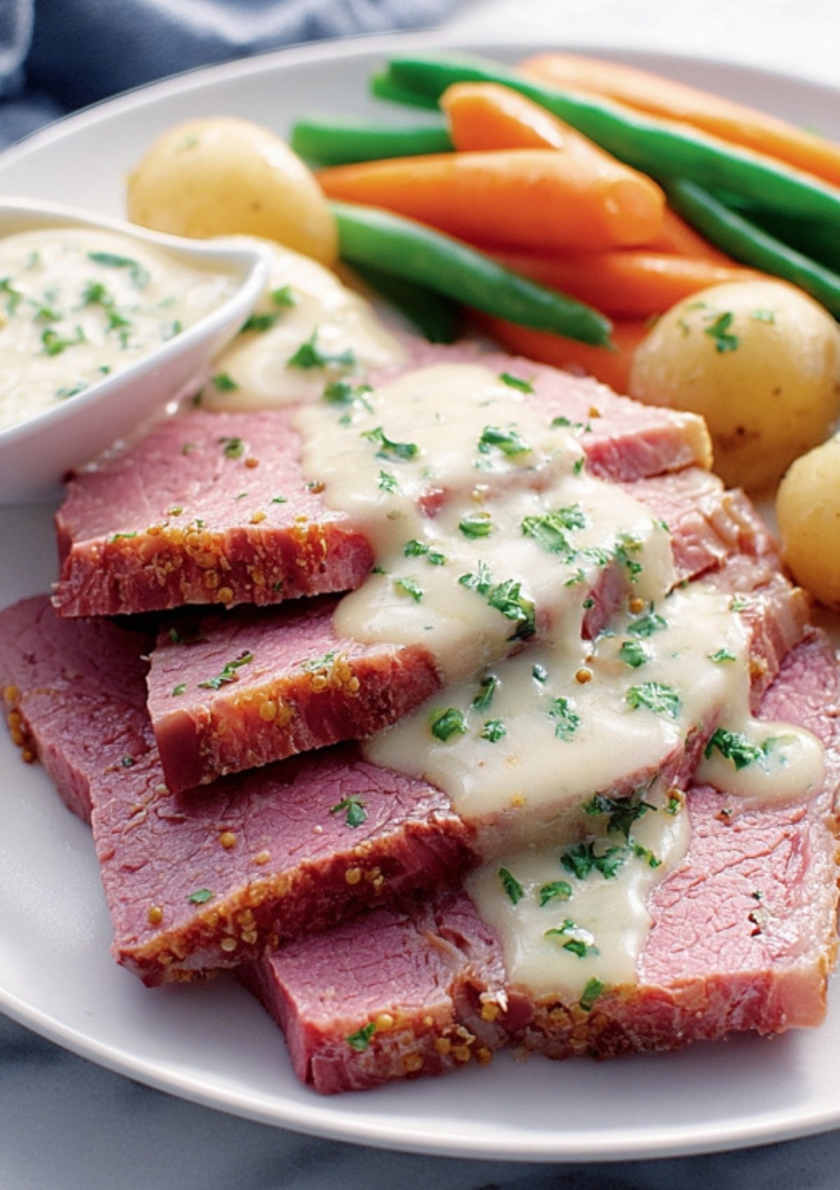 Sliced corned beef topped with creamy parsley sauce, served with baby potatoes, carrots, and green beans on a white plate.