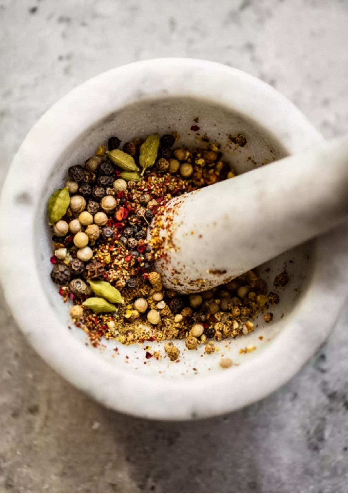Whole pickling spices including peppercorns, coriander, and cardamom being lightly crushed in a marble mortar and pestle for homemade corned beef.